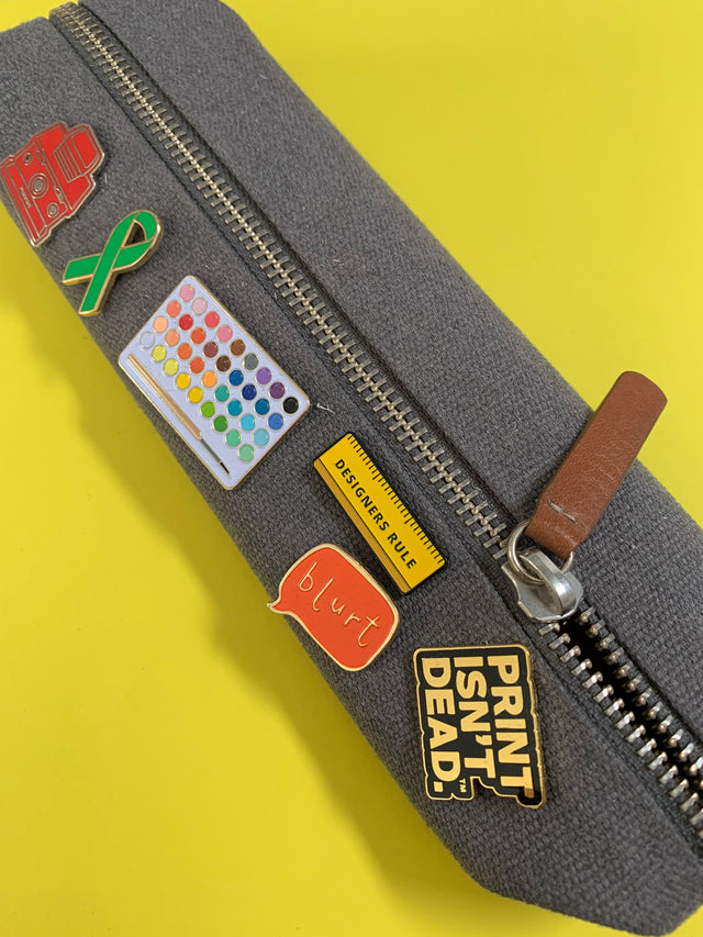 A pencil case with a variety of enamel pins stuck on it. A green ribbon, a painters palette, a mini yellow ruler, one saying print is not dead